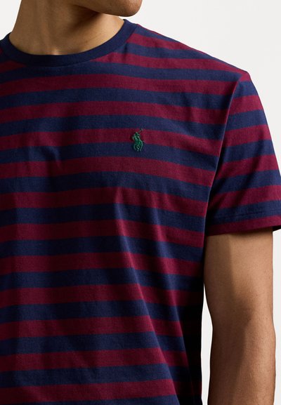 Rdeča in mornarsko modra črtasta majica iz bombaža s krožnim izrezom in zelenim vezenim logotipom na prsih. Kratki rokavi z sproščenim krojem.