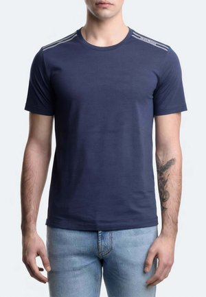 Uomo che indossa una t-shirt blu scuro a maniche corte con strisce chiare sulle spalle e jeans azzurri, in piedi con le braccia rilassate.