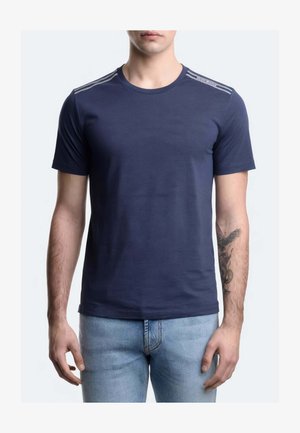 Uomo che indossa una t-shirt blu scuro a maniche corte con strisce chiare sulle spalle e jeans azzurri, in piedi con le braccia rilassate.