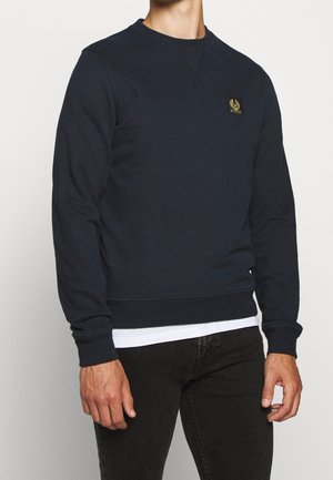 Man draagt een zwart, lang mouw sweatshirt met een klein logo op de borst, gecombineerd met een wit shirt en zwarte broek.