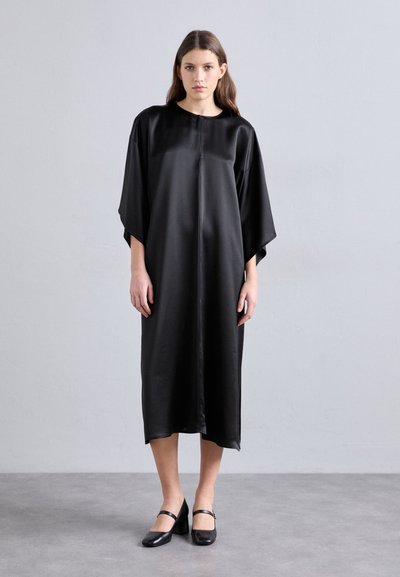 Vestido de satén negro con un corte relajado, que presenta un escote redondo y mangas murciélago. Combinado con zapatos de charol negros de estilo Mary Jane con tacón.