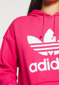 Primer plano de una persona vistiendo una sudadera Adidas de un brillante color rosa con el logo trefoil blanco y cordones, contra un fondo liso.