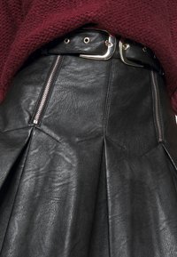 Černá kožená sukně s sklady, s viditelnými zipy a širokým páskem se stříbrnými detaily. Textura se zdá být hladká a strukturovaná.