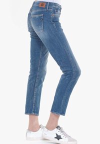 Vaqueros de denim en azul claro con un corte ajustado, que cuentan con bolsillos traseros y un parche de cuero en la cintura. Combinados con zapatillas blancas.