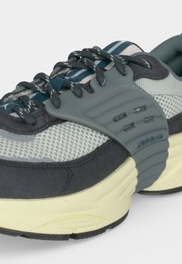 Παπούτσι τύπου sneaker με χαμηλή σιλουέτα, πάνω μέρος από γκρι διχτυωτό ύφασμα, επενδύσεις από σκούρο γκρι σουέτ, χαρακτηριστικό γκρι πλαστικό κλωβό για τα κορδόνια, μεσαία σόλα σε κρεμ απόχρωση και κορδόνια με σχέδια.