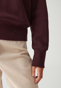 Mano appoggiata al fianco, indossa pantaloni in corduroy rigati beige e un top a maniche lunghe bordeaux con polsini ampi.