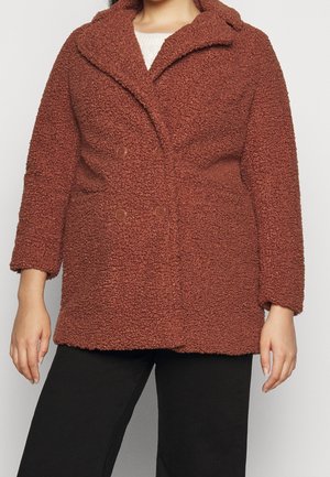 Femme portant un manteau marron texturé à double boutonnage avec de larges revers et un pantalon noir, debout sur un fond uni.