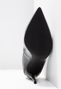 Chaussure noire à talon haut avec un bout pointu, tige en cuir brillant et talon stiletto fin ; semelle extérieure texturée noire et bordure argentée étincelante.