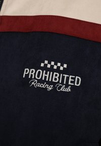 Tissu sombre avec texte brodé blanc "PROHIBITED Racing Club" sous un motif de drapeau de course à damiers blanc, bordé d'une bande rouge.