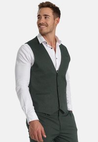 Mørkegrønn fiskebeinvest med tre knapper, hakkede slag og en enkelt brystlomme, båret over en hvit skjorte.