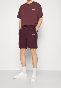 Homme portant un t-shirt et un short assortis Les Deux de couleur violet foncé, avec des chaussettes blanches et des baskets blanches, debout devant un fond clair uni.