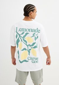 Camiseta blanca de gran tamaño con un gráfico de limón en verde y amarillo que incluye el texto "Lemonade" y "Citrus God" en la parte posterior. Mangas cortas.