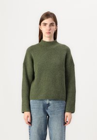 Donkergroene gebreide trui met een hoge halslijn, een relaxte pasvorm en lange mouwen, gecombineerd met lichtblauwe denimjeans.