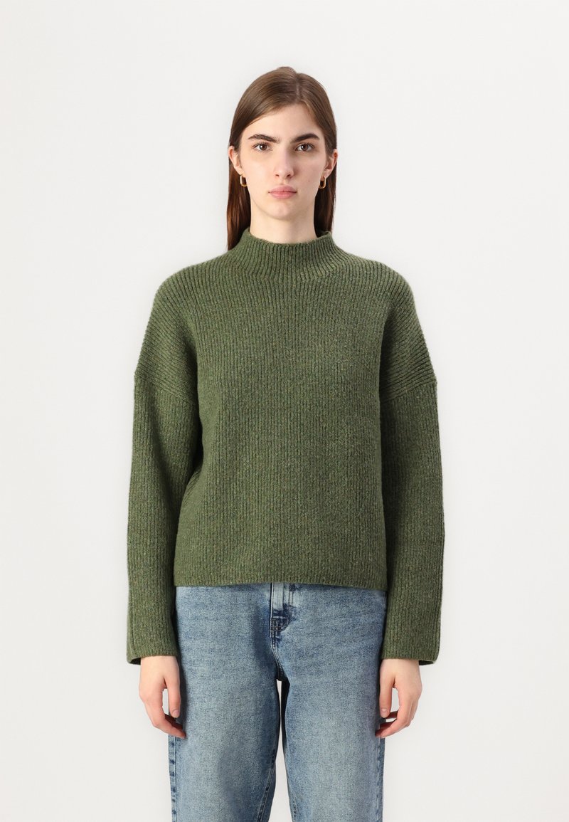 Donkergroene gebreide trui met een hoge halslijn, een relaxte pasvorm en lange mouwen, gecombineerd met lichtblauwe denimjeans.