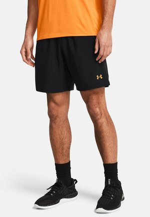Mężczyzna w czarnych sportowych szortach z pomarańczowym logo, pomarańczowej koszulce, czarnych skarpetach i czarnych butach do biegania stojący na białym tle.