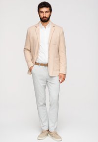 Homme avec une barbe portant un blazer beige, une chemise blanche, un pantalon gris clair, une ceinture marron et des chaussures beiges, debout avec une main dans la poche.