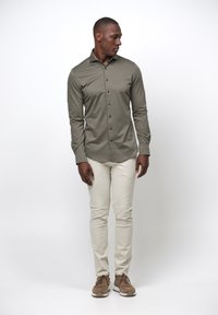 Camicia bottone verde oliva chiaro con colletto strutturato, maniche lunghe e bottoni neri, abbinata a pantaloni beige attillati e sneakers marroni.