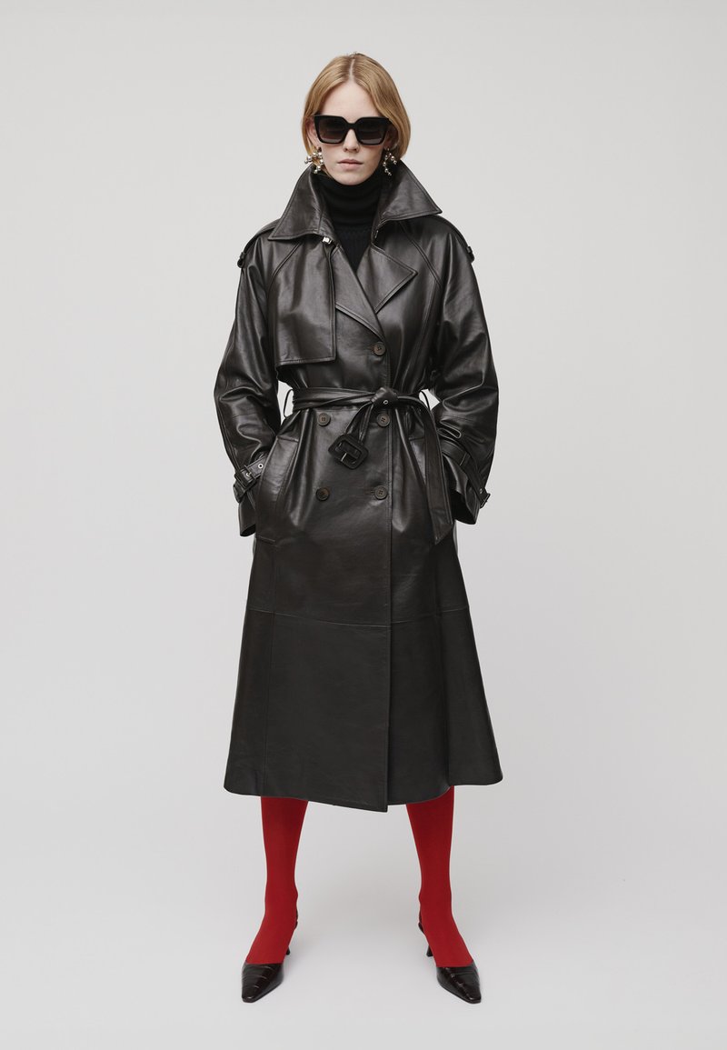 Frau in einem schwarzen Leder-Trenchcoat, schwarzem Rollkragenpullover, roten Strumpfhosen, schwarzen spitzen Schuhen, übergroßen schwarzen Sonnenbrille und großen Ohrringen.