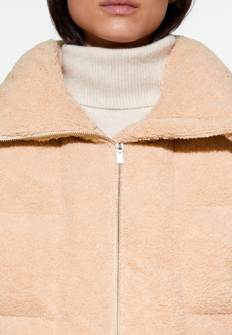 Pfirsichfarbene, flauschige Zip-Jacke mit Stehkragen und einem strukturierten, gesteppten Stoff. Darunter wird ein cremfarbener, gerippter Rollkragenpullover getragen.