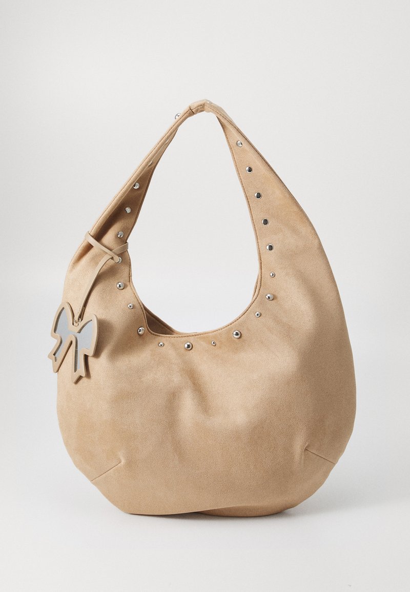 Beige semsket hobo-veske med sølvnagger langs toppen og en dekorativ sløyfe som aksent. Myk tekstur og rund form.