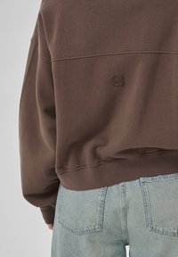 Brown sweatshirt with a relaxed fit, featuring a small embossed logo on the back. Paired with light blue high-waisted jeans.