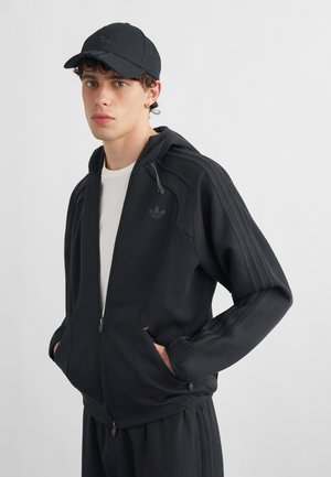 Ung mand iført en sort Adidas hættetrøje med lynlås, sort kasket, hvid skjorte og sorte bukser, stående med hænderne i lommerne mod en ensfarvet baggrund.
