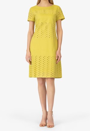 Femme portant une robe jaune mi-longue avec des manches courtes et des motifs en dentelle ajourée sur les manches, la poitrine et l'ourlet, assortie à des talons nus avec bride à la cheville.