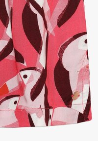 Odważny materiał z abstrakcyjnymi kształtami ptaków w odcieniach różu, czerwieni i bieli, z ciemnymi akcentami oraz małą okrągłą metalową zawieszką w pobliżu stębnowanego dołu.