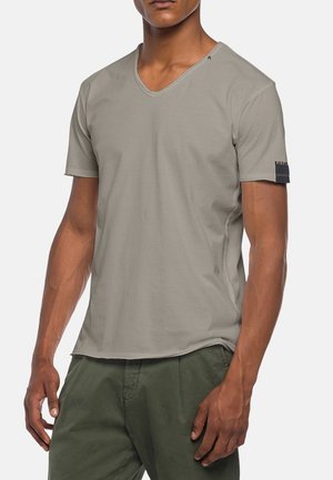 Homme portant un t-shirt gris clair à col en V et manches courtes, et un pantalon vert olive, debout les bras détendus le long du corps.
