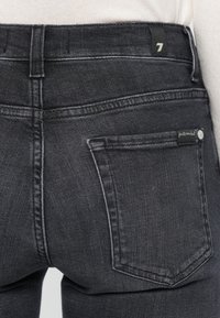 Mørkegrå denimjeans med en definert livlinje, baklommer med sømdetaljer og et merke sydd fast på livlinen.