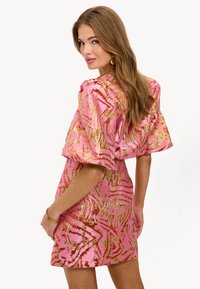 Vestido corto rosa con patrones abstractos metálicos dorados, mangas abullonadas y una silueta ajustada, que presenta una tela texturizada y un escote redondeado.