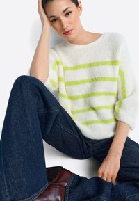 Pulover alb pufoș cu dungi verde lime, decolteu rotund și mâneci trei sferturi, asortat cu blugi din denim închis și cizme maro.