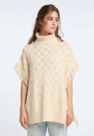 Crèmekleurige gebreide poncho met een hoge geribbelde kraag, ruitvormig kantpatroon, franjes en een ontspannen pasvorm, gecombineerd met lichte jeans.