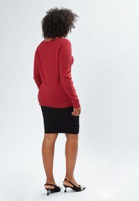 Maglione rosso a maglia con maniche lunghe, polsini e orlo ribattuti. Gonna a tubino nera con motivo testurizzato. Scarpa nera con tacco e decorazioni.