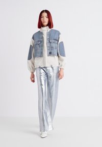 Denim- og fleecejakke med to forlommer, parret med skinnende sølvfarvede bukser med vide ben og hvide slip-on sko. Rødt hår står i kontrast til baggrunden.
