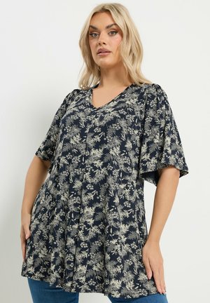 Lyshåret kvinde iført en løs, kortærmet navy tunika med lyse blomsterprint, stående foran en ensfarvet hvid baggrund.