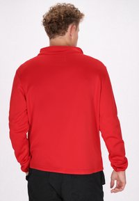 Rode lange mouwen polo met een kraag, gladde textuur en een subtiele gestikte detail op de achterkant van de nek. Draagt over donkere broeken.