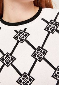 Gros plan sur un pull blanc avec des motifs géométriques en forme de losange noirs et un col rond côtelé noir.