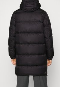 Svart pufferjacka med huva, med en quiltad design, tjock stoppning och elastiska manschetter. Baksidan är slät med en logodetalj i fållen.