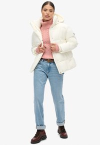 Chaqueta de plumas blanca con capucha, con un diseño acolchado y botones a presión, combinada con un suéter de punto trenzado rosa y jeans azul claros.