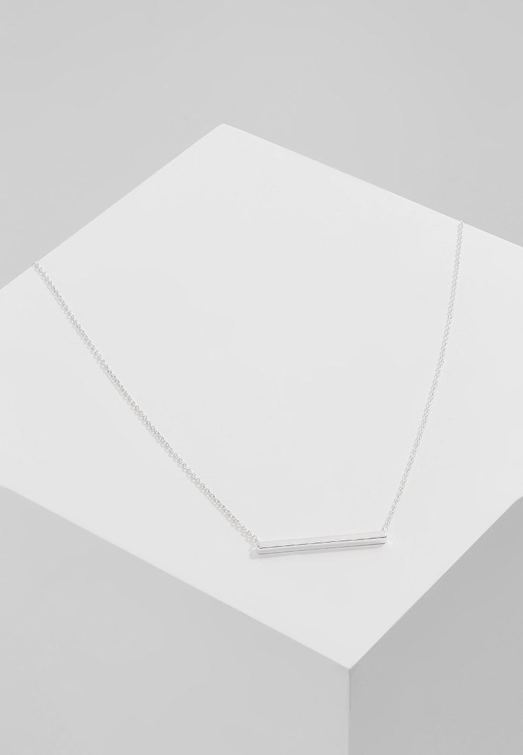 Collier en argent avec un pendentif barre fine horizontale présenté sur une surface géométrique blanche sur un fond gris clair.