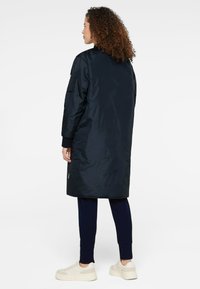 Langer, marineblauer Mantel aus glänzendem Material, mit Rippbündchen und einer Patttasche am linken Arm. Getragen mit dunklen Hosen und hellen Sneakers.