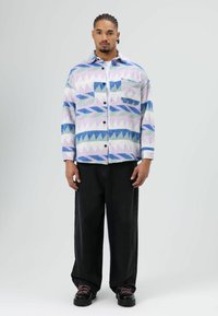 Multicolour vlinderhemd in blauw, paars en groen met geometrische patronen, zwarte knopen en een zak, gecombineerd met zwarte wijde broekspijpen.