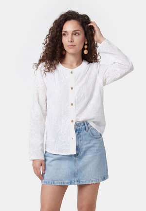 Kvinde med krøllet hår, iført runde hængeøreringe, en hvid broderet bluse med lange ærmer og et lyseblåt jeansskørt, stående mod en hvid baggrund.