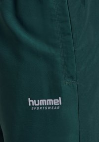 Gros plan sur un tissu de sport vert foncé avec le logo "hummel SPORTSWEAR" imprimé en gris clair près d'une couture.