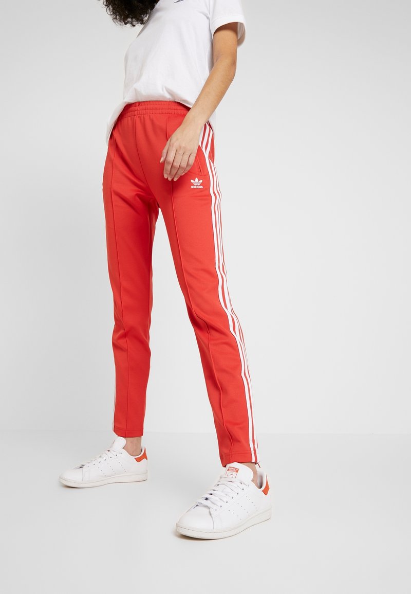 Červené sportovní kalhoty z hladkého materiálu s bílými bočními pruhy a logem Adidas na levém boku, doplněné o bílé tenisky.