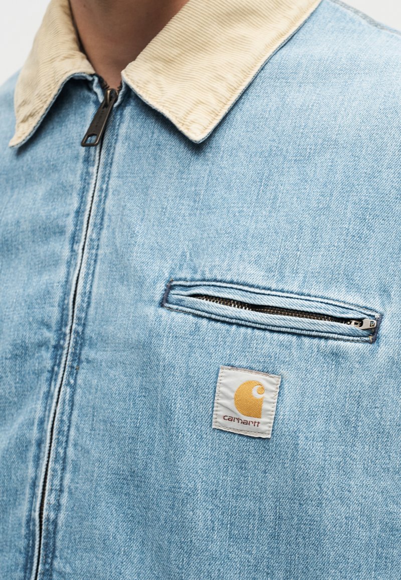 Lichtblauwe spijkerjas met beige ribfluwelen kraag, rits aan de voorkant, ritszak op de borst en Carhartt-logo op de borst.