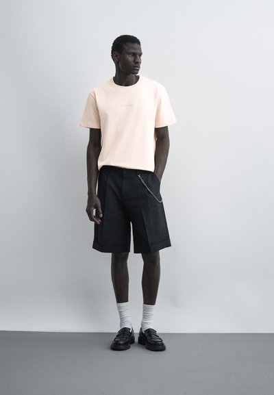 Άνδρας που στέκεται φορώντας ανοιχτό ροζ μπλουζάκι, μαύρα σορτς μέχρι το γόνατο με αλυσίδα, λευκές κάλτσες και μαύρα loafers σε μονόχρωμο γκρι φόντο.