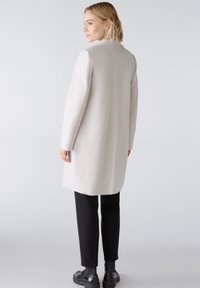 Hellbeige Mantel mit strukturiertem Finish, langen Ärmeln und taillierter Silhouette, kombiniert mit schwarzen Hosen und Schuhen.