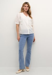 Camicia bianca con bottoni, motivo a occhio di pernice e colletto a volant, abbinata a jeans azzurri a zampa d'elefante e scarpe con tacco a blocco beige.